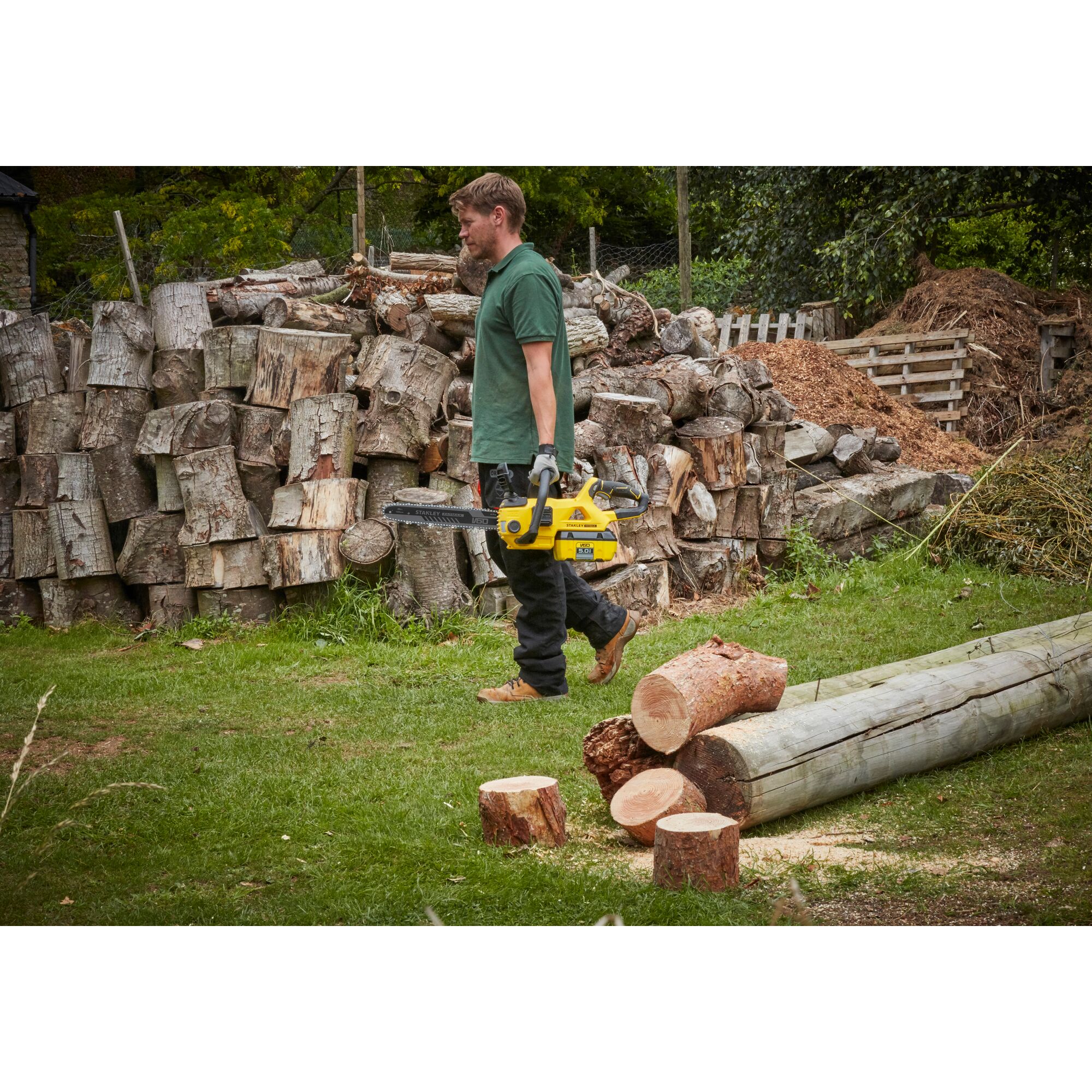 STANLEY SFMCCSB664B chainsaw held by person outdoors near cut logs and stacked firewood.